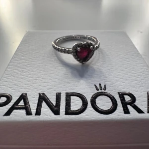 Silverring med hjärta från Pandora - Säljer en superfin silverring från Pandora med ett rött hjärta i mitten. Ringen är i storlek 48 och har en snygg, detaljerad design. Perfekt för att ge lite extra kärlek till din outfit!  Låda och påse ingår!!!