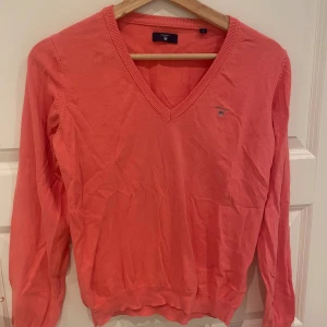 Korallfärgad tröja med V-ringning - Säljer en snygg korallfärgad tröja med V-ringning. Den är perfekt för både vår och höst och har långa ärmar. Tröjan är i bra skick och har en liten logga på bröstet. Perfekt för en casual look!