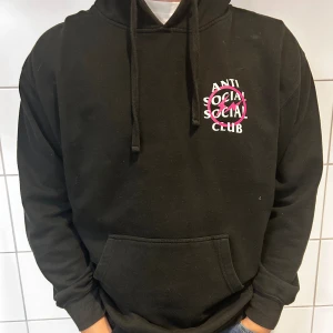 Svart hoodie från Anti Social Social Club - Säljer en svart hoodie från Anti Social Social Club. Den har en stor logga i vitt och rosa både fram och bak. Perfekt för en avslappnad stil och att hålla sig varm. Den har en klassisk känguruficka och justerbar huva med snören.