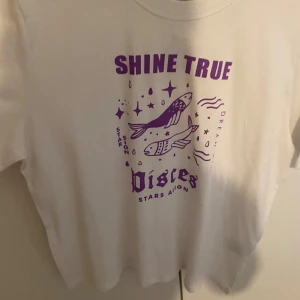 Vit t-shirt med lila tryck från Divided - Säljer en vit t-shirt från Divided med ett coolt lila tryck. Texten 'SHINE TRUE' och 'Pisces' tillsammans med stjärnor och en fiskdesign ger en unik look. Perfekt för en avslappnad stil! helt ny med prislapp på