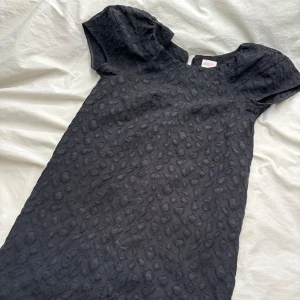 Svart cocktailklänning - En superfin cocktailklänning i ett svart tyg med mönster. Den har en dragkedja på baksidan av klänningen som är lång så den är lätt att ta på sig. Den går till strax över knäna.