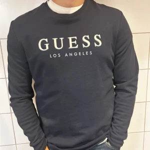Svart tröja från Guess - Säljer en stilren svart tröja från Guess med texten 'Guess Los Angeles' tryckt på framsidan. Tröjan har långa ärmar och en normal passform, perfekt för både vardag och fest. Superbekväm och lätt att matcha med olika outfits!
