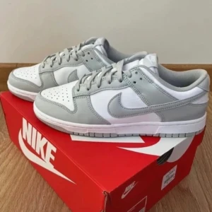 Nike Dunk Low i grått och vitt - Säljer ett par snygga Nike Dunk Low sneakers i en stilren kombination av grått och vitt. Skorna har en klassisk design med snörning och är perfekta för både vardag och fest. De är i nyskick och kommer i originalkartong.
