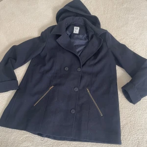 Svart rock från Crocker - Säljer en snygg svart rock från Crocker i storlek L. Den har en klassisk design med dubbelknäppning och två dragkedjefickor framtill. Perfekt för höst och vinter med sin stilrena look och praktiska huva. Rocken är i bra skick och redo för nya äventyr!