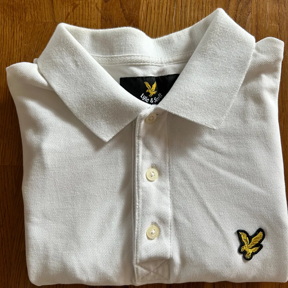 Säljer en snygg vit pikétröja från Lyle & Scott. Använd endast en gång. Storlek S. Tröjan har en klassisk krage med tre knappar och en broderad gul örnlogga på bröstet. . T-shirts.