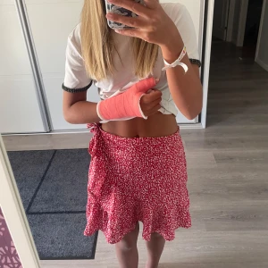 Röd omlottkjol med vita prickar - Säljer en superfin röd omlottkjol med vita prickar. Kjolen har en volangdetalj längst ner och knytning i midjan. Perfekt för sommaren eller en festlig tillställning. Den är i nyskick och passar perfekt till en avslappnad eller uppklädd stil. Materialet känns lätt och luftigt, vilket gör den bekväm att bära hela dagen.