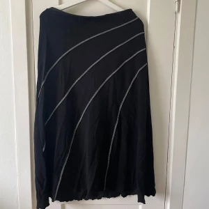 Svart långkjol med vita detaljer - Säljer en svart långkjol med vita diagonala linjer som ger en snygg och unik look. Kjolen är i ett mjukt och bekvämt material, perfekt för både vardag och fest. Den har en enkel design utan dragkedja eller knappar, vilket gör den lätt att ta på och av. Passar bra till alla säsonger och kan stylas på många olika sätt.