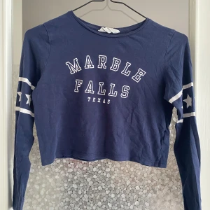 Blå croppad tröja med text - Säljer en blå croppad tröja med texten 'Marble Falls Texas' på framsidan. Tröjan har långa ärmar med vita ränder och stjärnor på ärmarna. Perfekt för en avslappnad och cool look! 💙