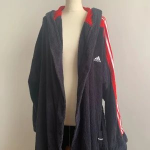 Svart och röd Adidas morgonrock - Säljer en supermysig svart och röd vintage Adidas morgonrock. Den har en huva och är gjord av mjukt fleece-material. Rocken har de klassiska Adidas-ränderna i vitt längs ärmarna och en stor Adidas-logga på ryggen. Perfekt för att hålla sig varm och bekväm hemma! pris kan diskuteras