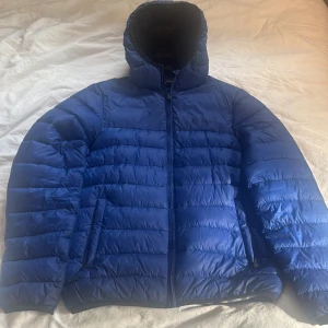 Blå dunjacka från Champion - Säljer en snygg blå dunjacka från Champion i storlek medium. Jackan har en quiltad design med dragkedja och en mysig huva. Perfekt för kalla vinterdagar när du vill hålla dig varm och stilren. Insidan är svart med Champion-loggan tryckt i vitt.
