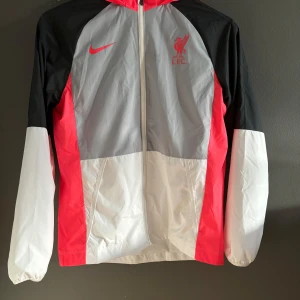 Liverpool jacka - Säljer en snygg och sportig vindjacka från Nike med LFC-logga. Jackan har en cool färgkombination av svart, grått, vitt och rött. Den är långärmad och har en dragkedja framtill. Perfekt för vår och höst! Nypris: 799