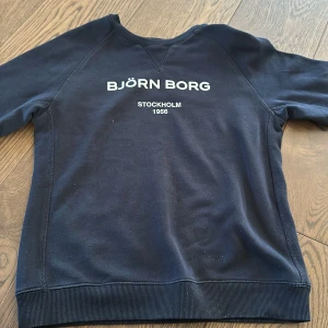 Mörkblå tröja från Björn Borg - Säljer en snygg och bekväm mörkblå tröja från Björn Borg. Den har ett stilrent tryck med texten 'BJÖRN BORG STOCKHOLM 1956' på framsidan. Tröjan är i bra skick och perfekt för både vardag och avslappnade tillfällen. Den är tillverkad i ett mjukt material som känns skönt mot huden.