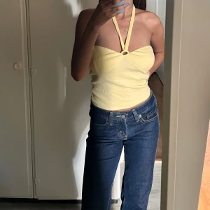 Gul halterneck-top  - Säljer en ljusgul halterneck-top med en elegant metallring som pryder bröstet och ger en sofistikerad touch. Denna topp är perfekt för sommardagar eller utekvällar och sitter skönt på kroppen tack vare stretchiga materialet som också framhäver midjan