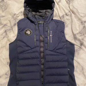 Mörkblå dunväst från Sail Racing - Säljer en snygg mörkblå dunväst från Sail Racing med huva och coolt märke på bröstet. Perfekt för kyliga dagar med sin quiltade design och dragkedja framtill. Västen är i mycket bra skick och passar perfekt för höst och vår. Den har en sportig look och är superbekväm att bära. 