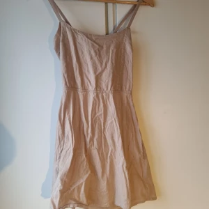 Beige klänning med knyt i ryggen - Snygg beige klänning med axelband och knytning i ryggen. Perfekt för varma dagar med sin lätta och luftiga design. Klänningen har en enkel och stilren look som passar till många tillfällen.