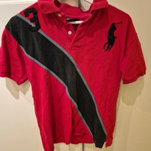 Röd pikétröja från Ralph Lauren - Snygg röd pikétröja från Ralph Lauren med korta ärmar och klassisk krage. Tröjan har en stor svart logotyp på bröstet och en diagonal rand i svart och grått. Perfekt för en sportig look! Står L men skulle säga M