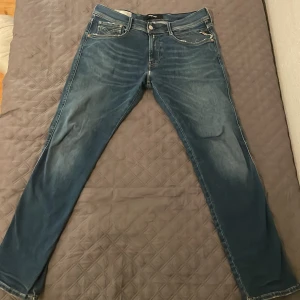 Blå jeans från Replay - Säljer ett par snygga blå jeans från Replay i modellen Anbass. De har en klassisk femficksdesign och en bekväm passform. Perfekta för både vardag och fest! 🕺hyperflex material. Otroligt sköna. Som ni ser så har de en slitning på framsidan men de har varit så ett tag så de ska vara lunga puckar. Byxorna är väl använda 