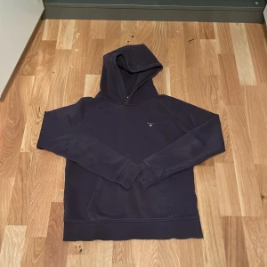GANT Hoodie  - Säljer en snygg mörkblå hoodie från GANT med broderad logga på bröstet. Tröjan har en klassisk design med huva och långa ärmar. Perfekt för en avslappnad stil.
