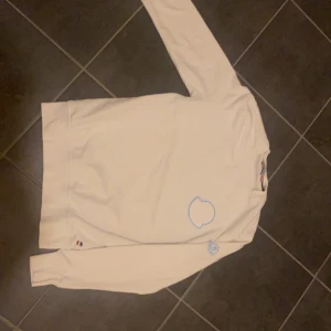 Vit tröja från Moncler - Säljer en stilren vit tröja från Moncler med deras ikoniska logga i blått på bröstet och ärmen. Tröjan har långa ärmar och är perfekt för en casual look. Passar både till vardags och lite finare tillfällen. Superbekväm och i bra skick!