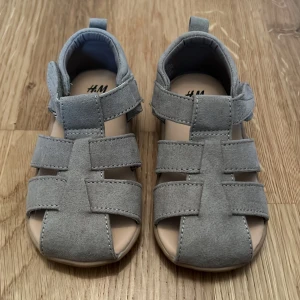Grå sandaler från H&M - Säljer ett par grå sandaler från H&M i storlek 22. Sandalerna har en praktisk kardborreknäppning och en bekväm sula för små fötter. Perfekta för varma sommardagar.