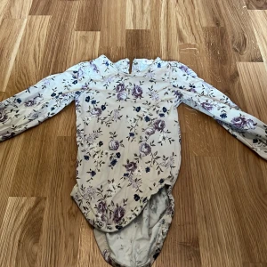 Blommig långärmad body från Newbie - Säljer en söt långärmad body från Newbie med ett vackert blommönster i lila och blått på en vit bakgrund. Perfekt för små barn i storlek 86 (12-18 månader).