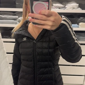 Svart pufferjacka från Adidas - Säljer en snygg svart pufferjacka från Adidas i storlek S. Jackan har långa ärmar med de klassiska tre ränderna längs ärmarna och en dragkedja framtill. Perfekt för höst och vinter, håller dig varm och stilren. 🖤