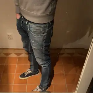 Säljer ett par snygga blå jeans med coola slitningar. De har en klassisk femficksdesign och är i ett bekvämt material som passar perfekt till vardags. Jeansen har en smal passform och är i bra skick med lite slitage för en trendig look. 