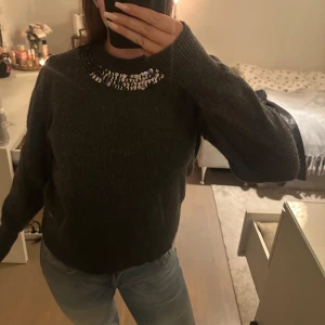 Mörkgrå stickad tröja med paljettdetaljer - Säljer en mysig mörkgrå stickad tröja med glittrande paljetter runt halsringningen. Perfekt för att ge lite extra bling till din outfit. Tröjan har långa ärmar och en bekväm passform.