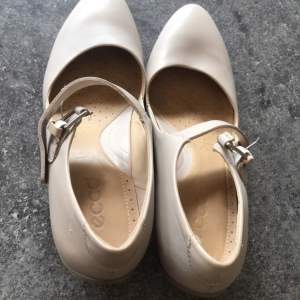 Snygga beige pumps från Ecco med en elegant rem och spänne över foten. Skorna har en bekväm klack och rundad tå, perfekt för en stilren look.