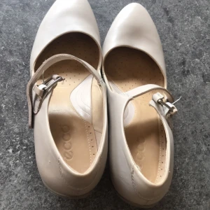 Beige pumps från Ecco - Snygga beige pumps från Ecco med en elegant rem och spänne över foten. Skorna har en bekväm klack och rundad tå, perfekt för en stilren look.