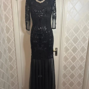 Svart långklänning med paljetter - Elegant svart långklänning med glittrande paljetter och spetsdetaljer. Klänningen har trekvartsärmar och en vacker broderad design. Perfekt för en glamorös kväll.