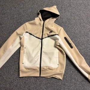Beige och vit huvtröja från Nike - Snygg och sportig huvtröja från Nike i beige och vitt. Tröjan har en dragkedja framtill och en praktisk ficka på ärmen. Perfekt för en avslappnad stil med sin bekväma passform och huva.