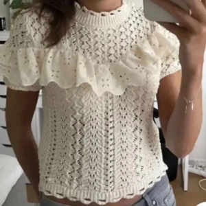 Vit stickad topp med volang - Charmig vit stickad topp med korta ärmar och volangdetalj över bröstet. Den har ett vackert hålmönster och ribbade kanter vid ärmslut och nederkant. Perfekt för en söt och stilren look.