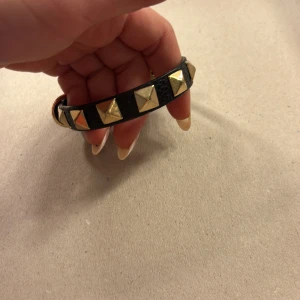 Svart armband med nitar - Snyggt svart armband med guldiga pyramidformade nitar. Armbandet har en justerbar rem med ett guldfärgat spänne för perfekt passform. Perfekt accessoar för att ge din outfit en edgy touch!
