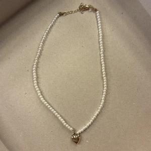 Pärlhalsband med hjärthänge - Säljer ett elegant pärlhalsband med ett guldigt hjärthänge. Halsbandet har små vita pärlor och en justerbar kedja för perfekt passform. Perfekt för att ge en romantisk touch till din outfit. Passar både till vardags och festliga tillfällen.
