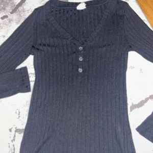 Svart ribbad tröja med knappar - Snygg svart ribbad tröja med v-ringning och tre knappar framtill. Tröjan har långa ärmar och en stilren design som passar perfekt till olika tillfällen. Väl använd.