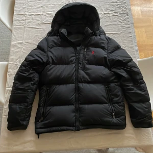 Svart dunjacka från Ralph Lauren - Säljer en snygg svart dunjacka från Ralph Lauren i mycket bra skick. Jackan har en praktisk dragkedja och en liten röd logga på bröstet. Perfekt för kalla vinterdagar med sin varma och quiltade design. Den har en huva och långa ärmar för extra skydd mot kylan. Passar både till vardags och lite finare tillfällen.