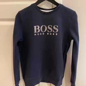 Mörkblå tröja från Hugo Boss - Säljer en snygg mörkblå tröja från Hugo Boss. Den har ett stilrent tryck med BOSS-loggan på framsidan. Tröjan är i bra skick och perfekt för både vardag och fest. Passar bra till jeans eller chinos för en avslappnad men ändå uppklädd look.