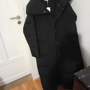 Svart lång dunjacka från VILA - Säljer en svart lång dunjacka från VILA i storlek 40. Jackan är perfekt för kalla vinterdagar med sin vadderade design och långa längd. Den har en hög krage och knappar framtill för extra värme. Ett måste för vintern! ❄️