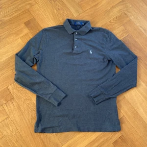 Grå pikétröja från Ralph Lauren - Säljer en stilren grå pikétröja från Ralph Lauren i storlek M. Tröjan har en klassisk krage med tre knappar och det ikoniska broderade logotypen på bröstet. Perfekt för både vardag och lite finare tillfällen. Passformen är custom slim fit, vilket ger en snygg siluett. 🐎