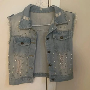 Jeansväst med pärlor - Snygg jeansväst med slitna detaljer och nitar. Västen har knappar framtill och två bröstfickor. Perfekt för en cool och avslappnad stil. Liten i st