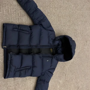 Mörkblå dunjacka från Polo Ralph Lauren - Säljer en snygg mörkblå dunjacka från Polo Ralph Lauren. Jackan har en praktisk huva och dragkedja framtill. Perfekt för kyliga dagar med sin vadderade design.