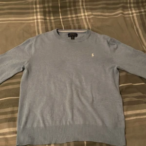 Ljusblå tröja från Polo Ralph Lauren - Säljer en stilren ljusblå tröja från Polo Ralph Lauren med det klassiska broderade logotypen på bröstet. Tröjan har rund hals och ribbade muddar vid ärmslut och nederkant. Perfekt för en avslappnad och snygg look. Liten defekt på bröstet bara.