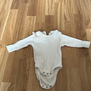 Vit ribbad body från Newbie - Söt vit ribbad body från Newbie i storlek 62 för 2-4 månader. Den har långa ärmar och fina volangdetaljer vid axlarna. Perfekt för små äventyrare!
