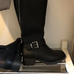 Svarta stövlar med dragkedja och spänne - Snygga svarta stövlar med dragkedja längs sidan och ett dekorativt spänne. Perfekta för höst och vinter, dessa stövlar har en robust sula och en stilren design.