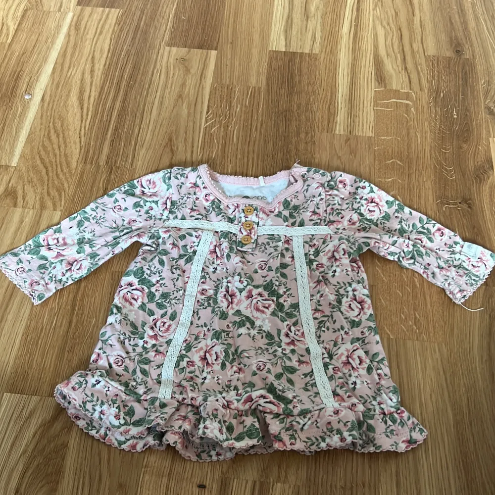 Söt blommig klänning i rosa och grönt från Tiny One. Tillverkad i ekologisk bomull med trekvartsärmar och volangdetaljer. Perfekt för små fashionistas!. Mekot.
