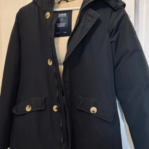 Svart parkajacka från Svea - Snygg svart parkajacka från Svea med huva och knappar i guldton. Jackan har praktiska fickor framtill och en broderad Svea-logga på bröstet. Perfekt för kyligare dagar.