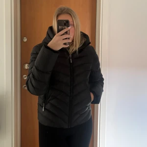 Svart pufferjacka från Chelsea - Snygg svart pufferjacka från Chelsea med dragkedja och huva. Perfekt för kyliga dagar med sin vadderade design. De står stl 42 men jag har stl M i mina kläder och jackor och denna sitter tight och är förliten så jag skulle säga att den är mer som än S