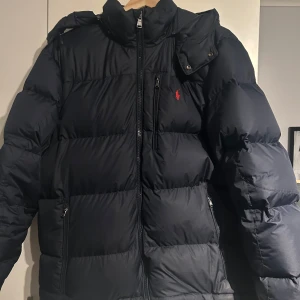 Mörkblå dunjacka från Polo Ralph Lauren - Säljer en mörkblå dunjacka från Polo Ralph Lauren i mycket bra skick. Jackan har en dragkedja framtill och är perfekt för vintern. Den är långärmad och har en klassisk pufferstil som håller dig varm och snygg. Perfekt för kalla dagar! 🖤