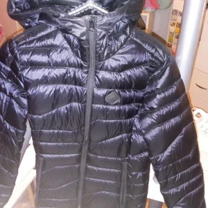 Svart pufferjacka från J.Lindeberg - Säljer en snygg svart pufferjacka från J.Lindeberg i storlek XS. Jackan har en glansig finish och är perfekt för kyliga dagar. Den har en dragkedja framtill och en mysig huva. Passar perfekt för både höst och vinter!